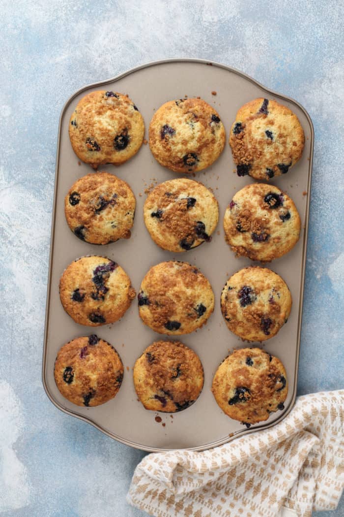 Freshly baked pan of blueberry crumb muffins set on a light blue countertop.