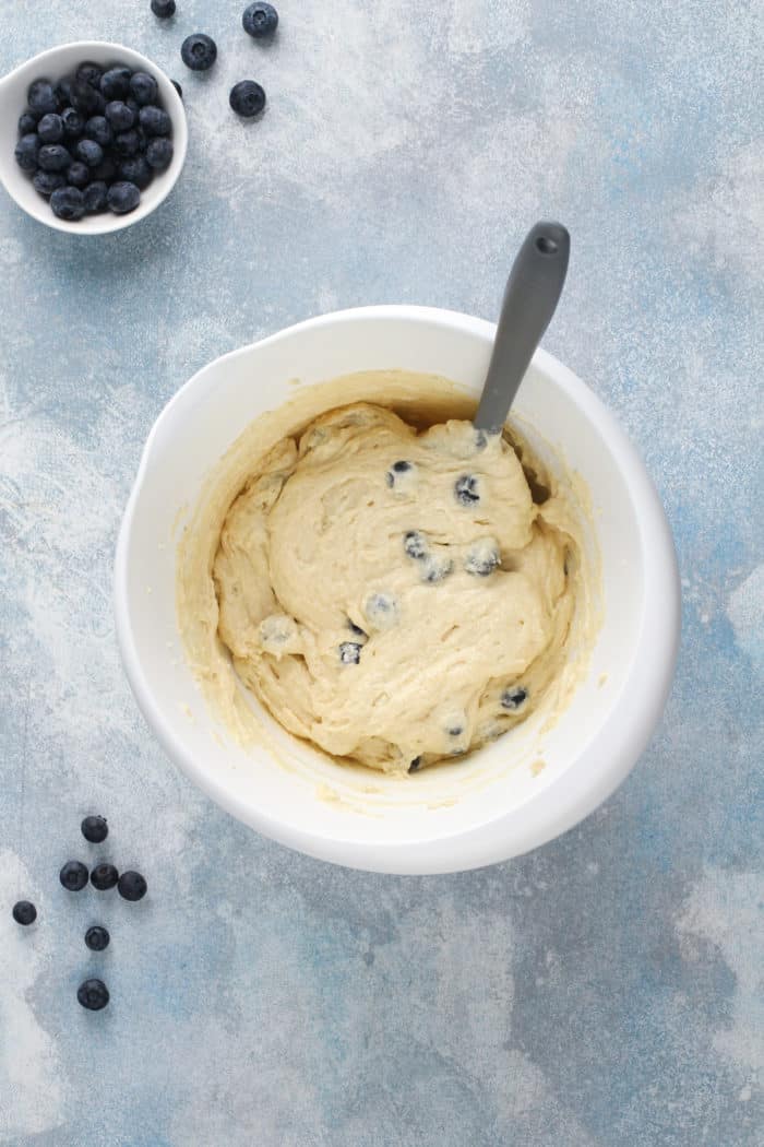 Blueberry muffin batter being stirred with a spatula in a white mixing bowl.