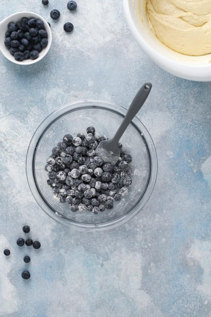 Blueberries tossed with flour in a glass mixing bowl, set on a blue countertop.