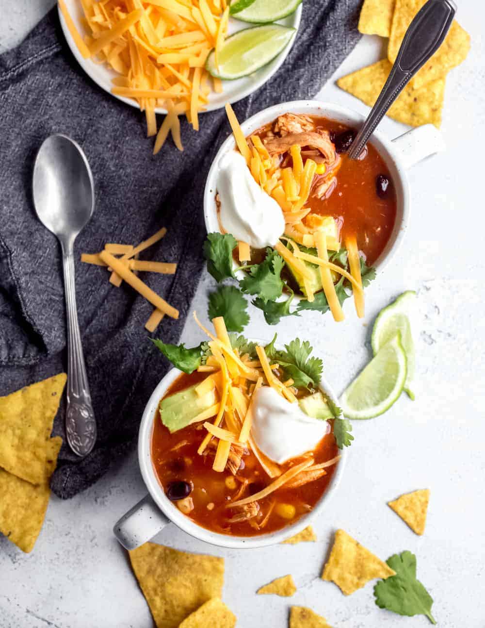 Two white mugs filled with tortilla soup and topped with cheese, avocado, sour cream and cilantro