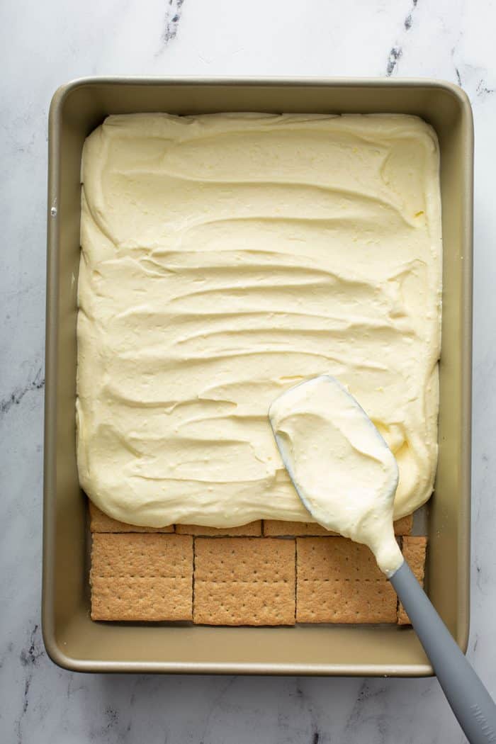 Spatula spreading vanilla filling on top of graham crackers in a baking pan for eclair cake