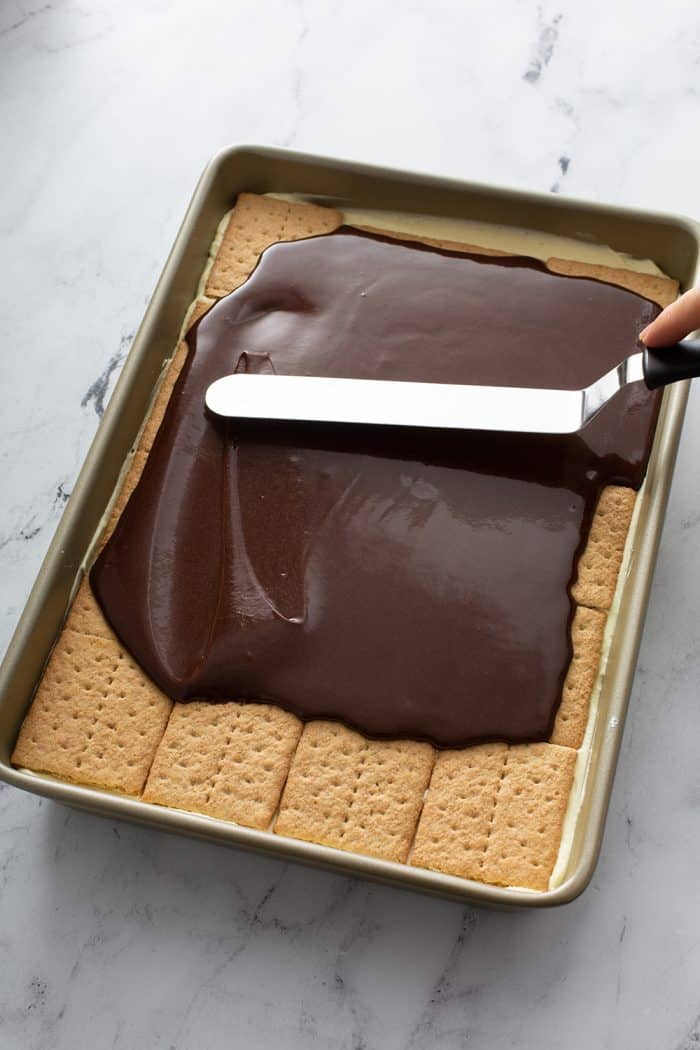 Offset spatula spreading chocolate frosting on top of eclair cake in a metal baking pan