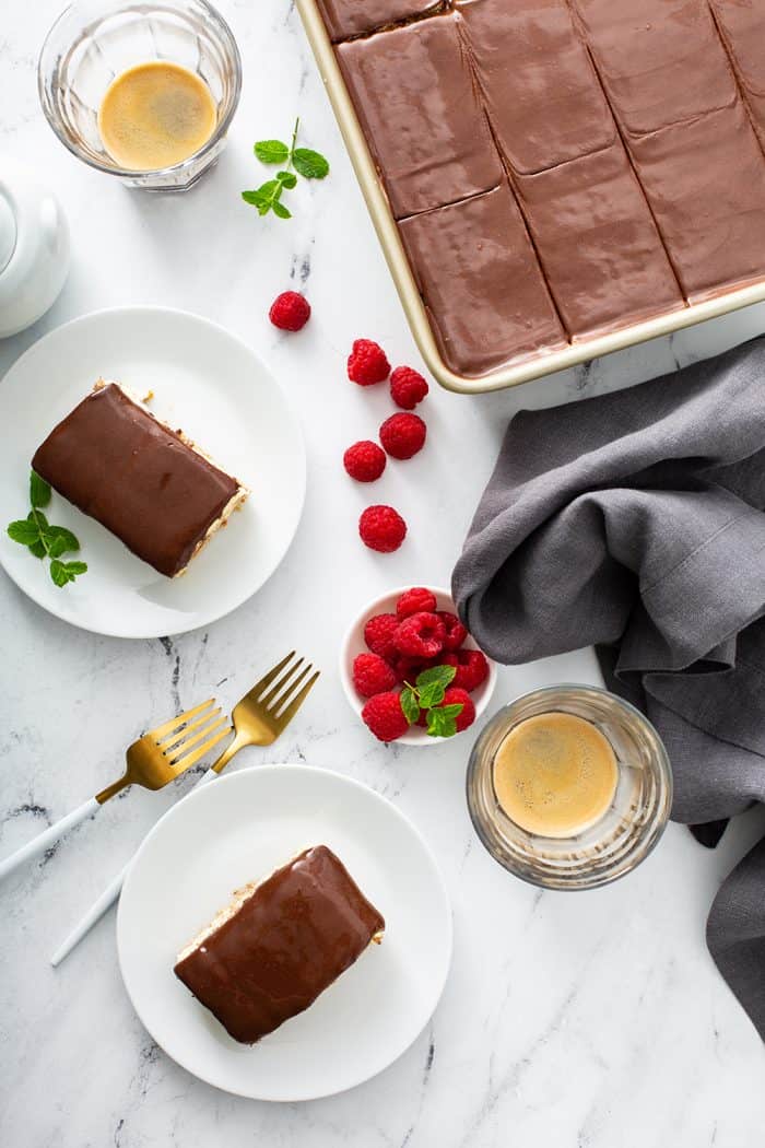 Overhead view of two white plates holding slices of eclair cake and a baking pan of eclair cake on a white counter scatter with fresh raspberries