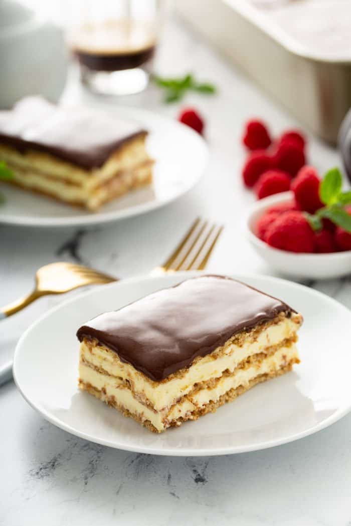 Slice of eclair cake on a white plate with another plate and fresh raspberries in the background