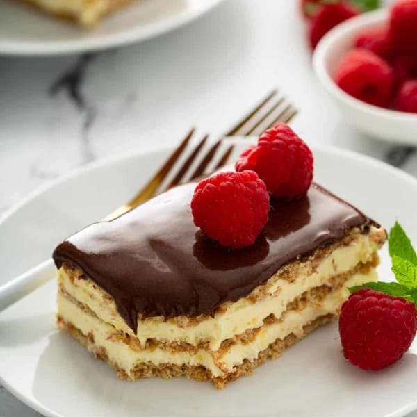 Fork next to a slice of eclair cake topped with fresh raspberries on a white plate
