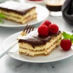 Slice of eclair cake topped with fresh raspberries on a white plate with a second slice of cake in the background