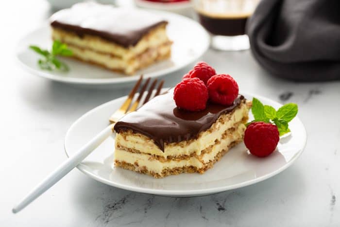 Slice of eclair cake topped with fresh raspberries on a white plate with a second slice of cake in the background