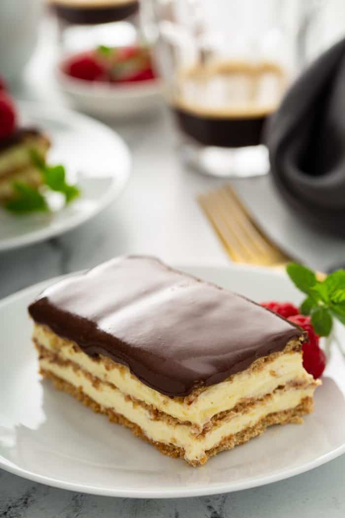 Slice of eclair cake on a white plate with cups of coffee and more plates in the background