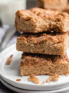 Three stacked snickerdoodle blondies on a white plate. The top blondie has a bite taken out of it.