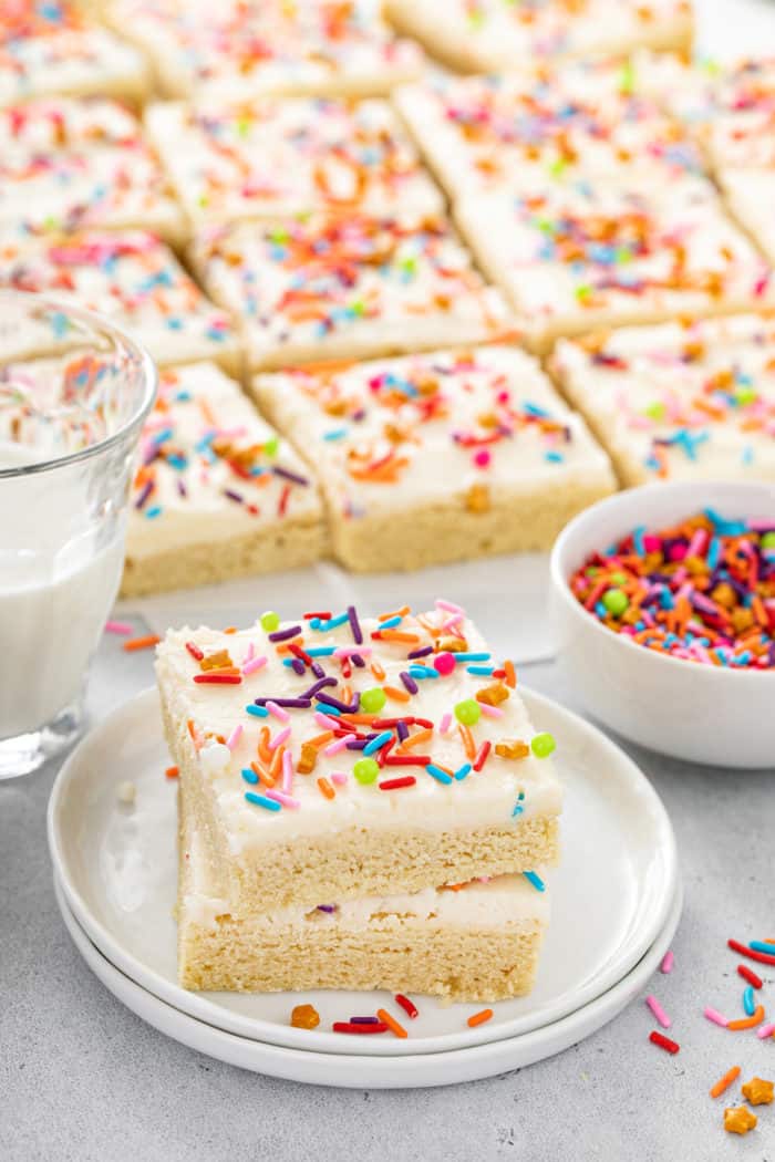 Two sugar cookie bars stacked on a white plate with more bars in the background