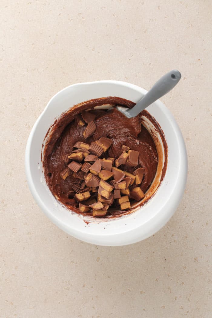 Chopped peanut butter cup pieces being folded into brownie batter in a white mixing bowl.