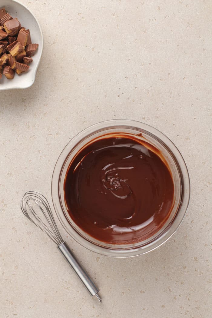 Melted chocolate in a glass mixing bowl, set next to a whisk.