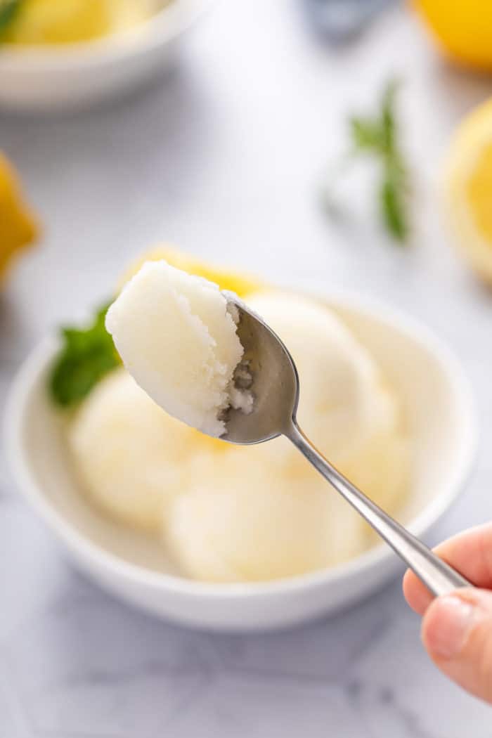 Spoon holding up a bite of lemon sorbet. A bowl of the sorbet is visible in the background.