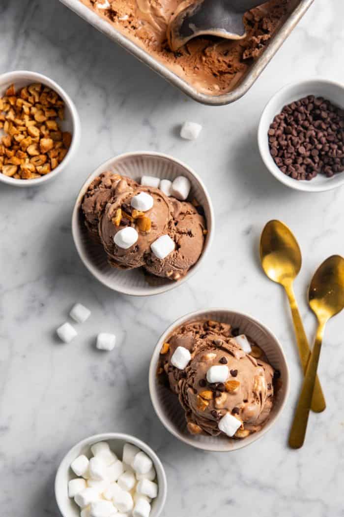 Two white bowls filled with rocky road ice cream and surrounded with bowls of nuts, chocolate chips, and mini marshmallows.