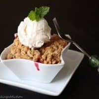 Cherry Crisp with a scoop of vanilla ice cream in a white, square bowl on a white square plate with a fork leaning on the right side of the bowl.