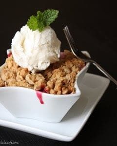 Cherry Crisp with a scoop of vanilla ice cream in a white, square bowl on a white square plate with a fork leaning on the right side of the bowl.
