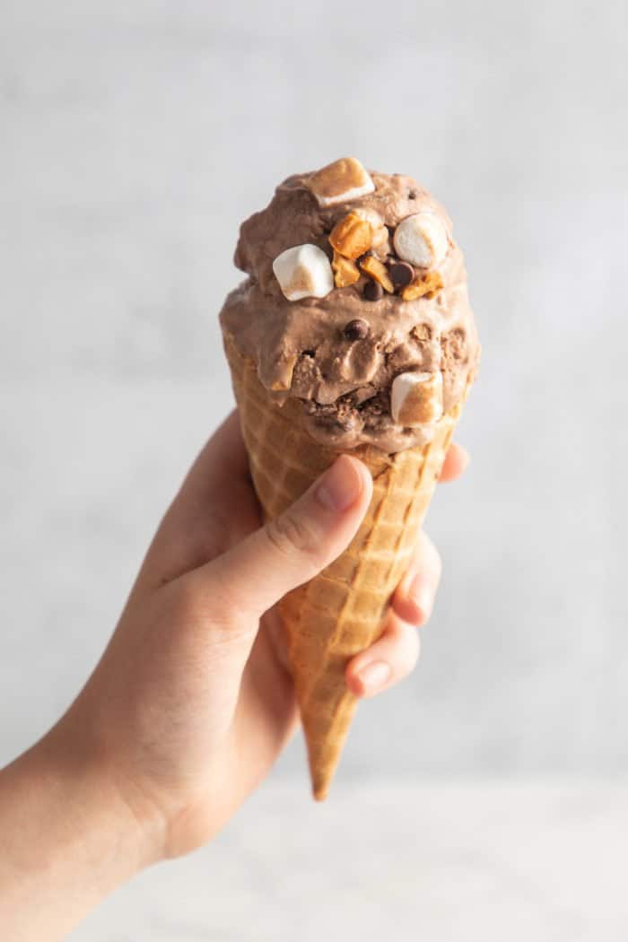 Hand holding up a waffle cone filled with rocky road ice cream in front of a light gray wall.
