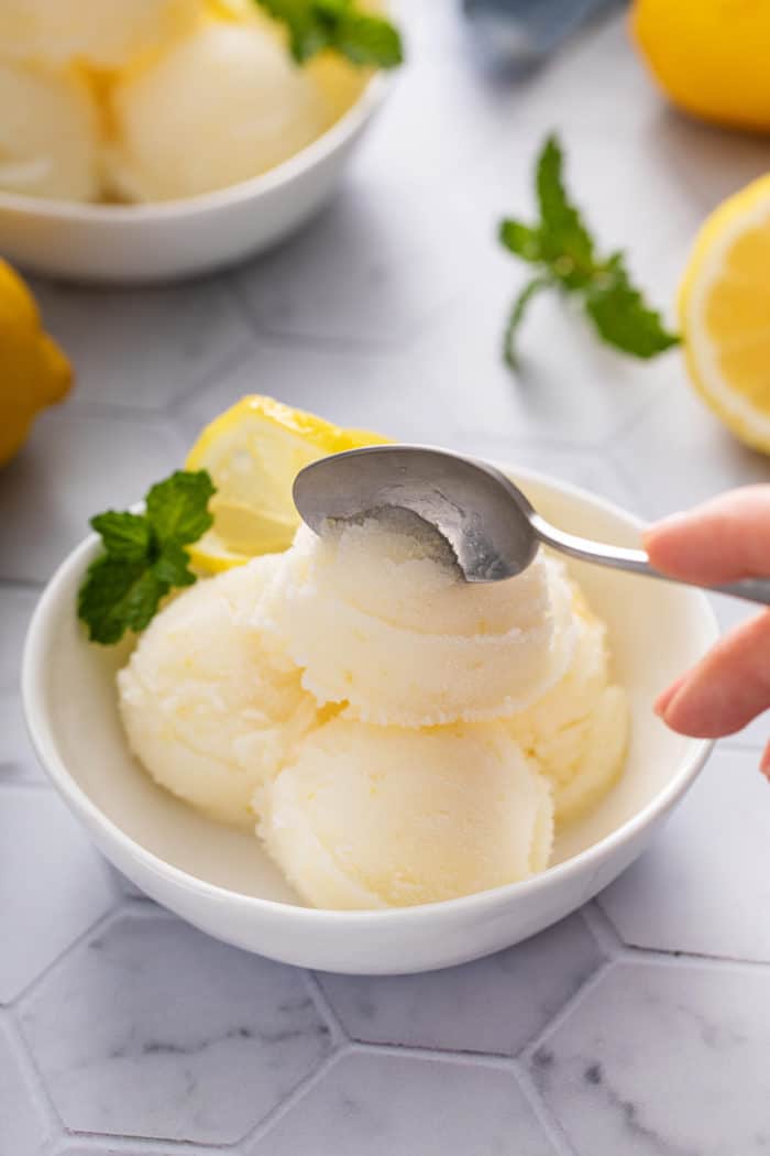 Hand dipping a spoon into a scoop of lemon sorbet in a white bowl.