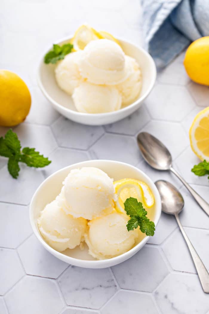 Two white bowls, each filled with several scoops of lemon sorbet, set on a white tile countertop.