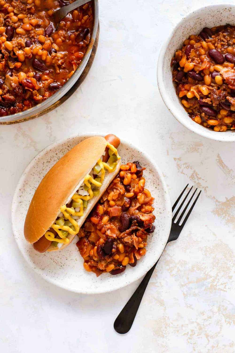 Hot dog and old fashioned baked beans on a white plate next to bowls of baked beans