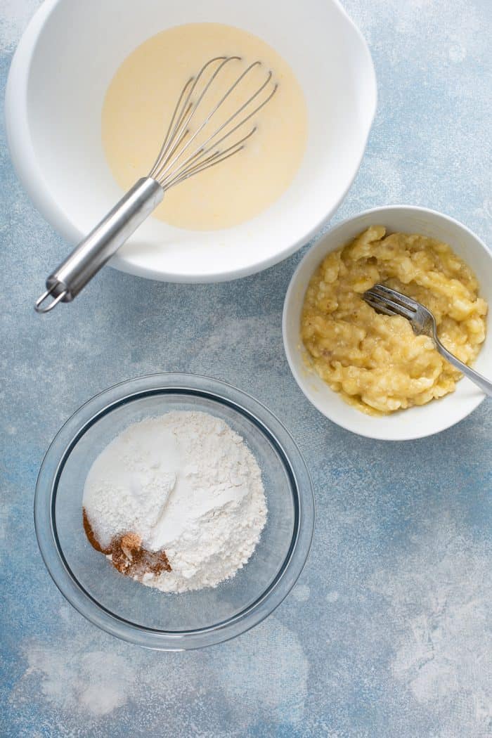 bowls containing the dry ingredients, wet ingredients, and mashed bananas for banana pancakes on a blue countertop