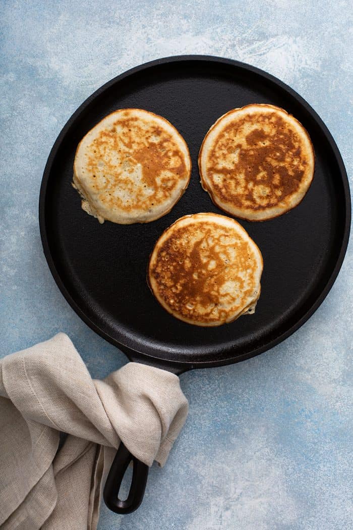 Griddle with 3 banana pancakes on a blue countertop, with a towel wrapped around the handle of the skillet