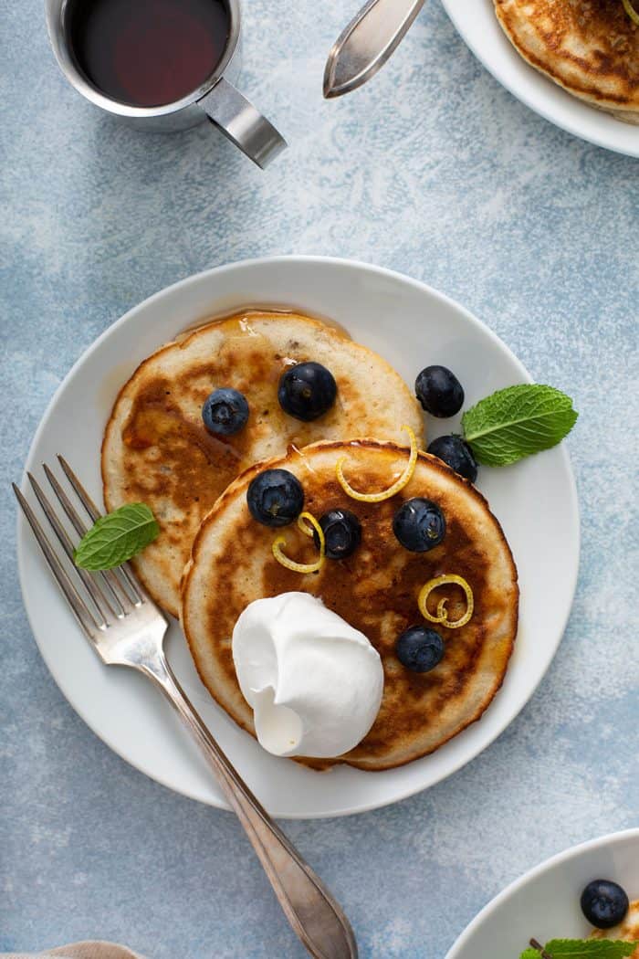 Two banana pancakes topped with blueberries, whipped cream and mint on a white plate set on a light blue surface