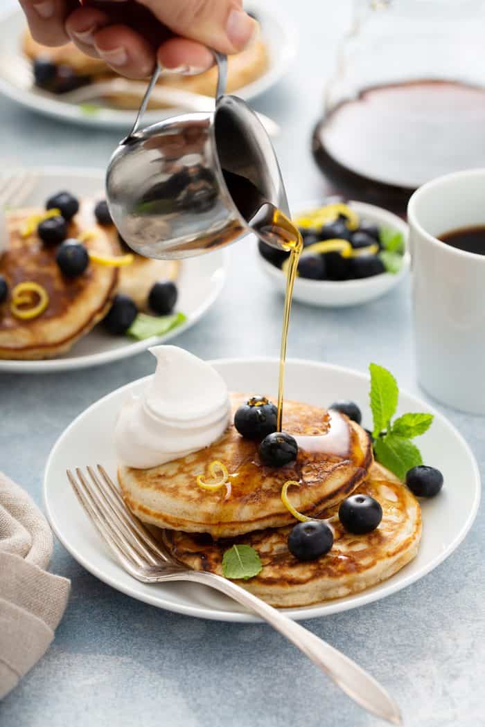 Syrup being poured over 2 banana pancakes topped with blueberries and whipped cream on a white plate. Two more plates of pancakes and a mug of coffee are in the background