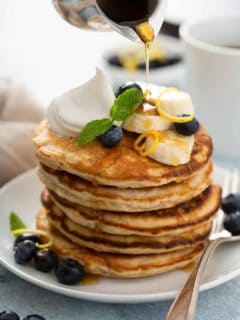Stack of banana pancakes on a white plate, topped with sliced bananas, blueberries and whipped cream, with syrup being drizzled on top