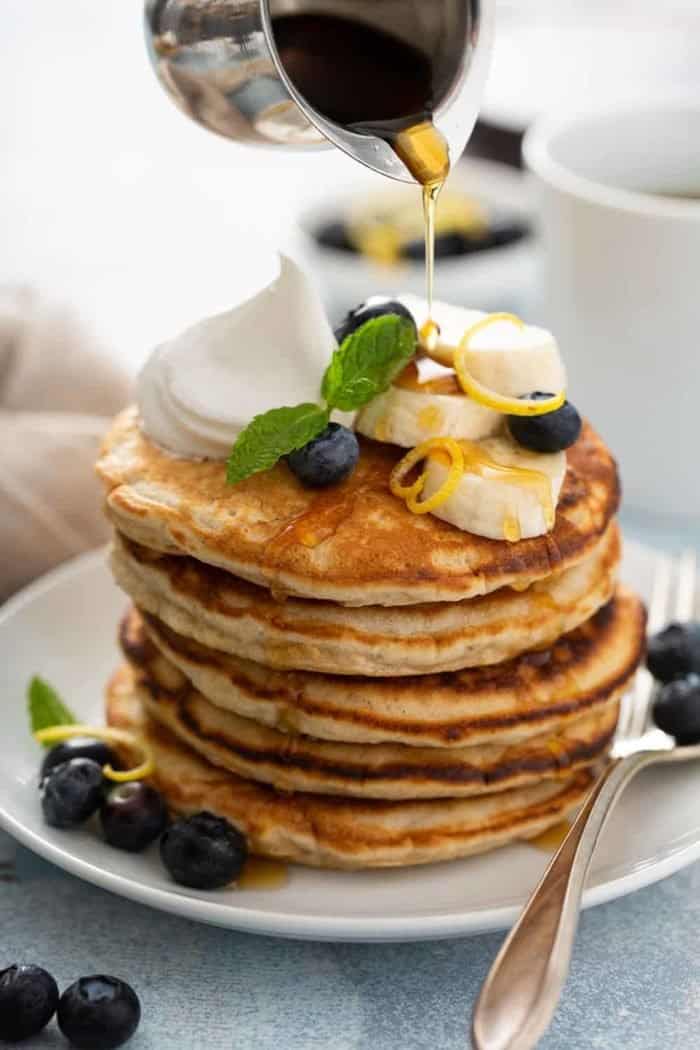 Stack of banana pancakes on a white plate, topped with sliced bananas, blueberries and whipped cream, with syrup being drizzled on top