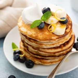 Stack of banana pancakes topped with blueberries, sliced bananas and whipped cream on a white plate, next to a fork