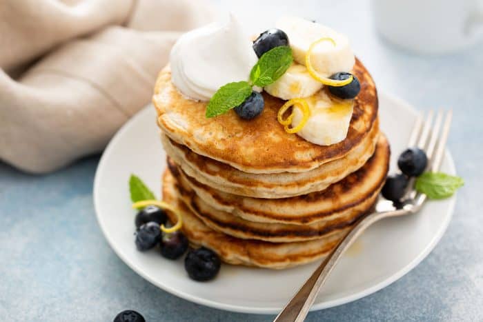 Stack of banana pancakes topped with blueberries, sliced bananas and whipped cream on a white plate, next to a fork