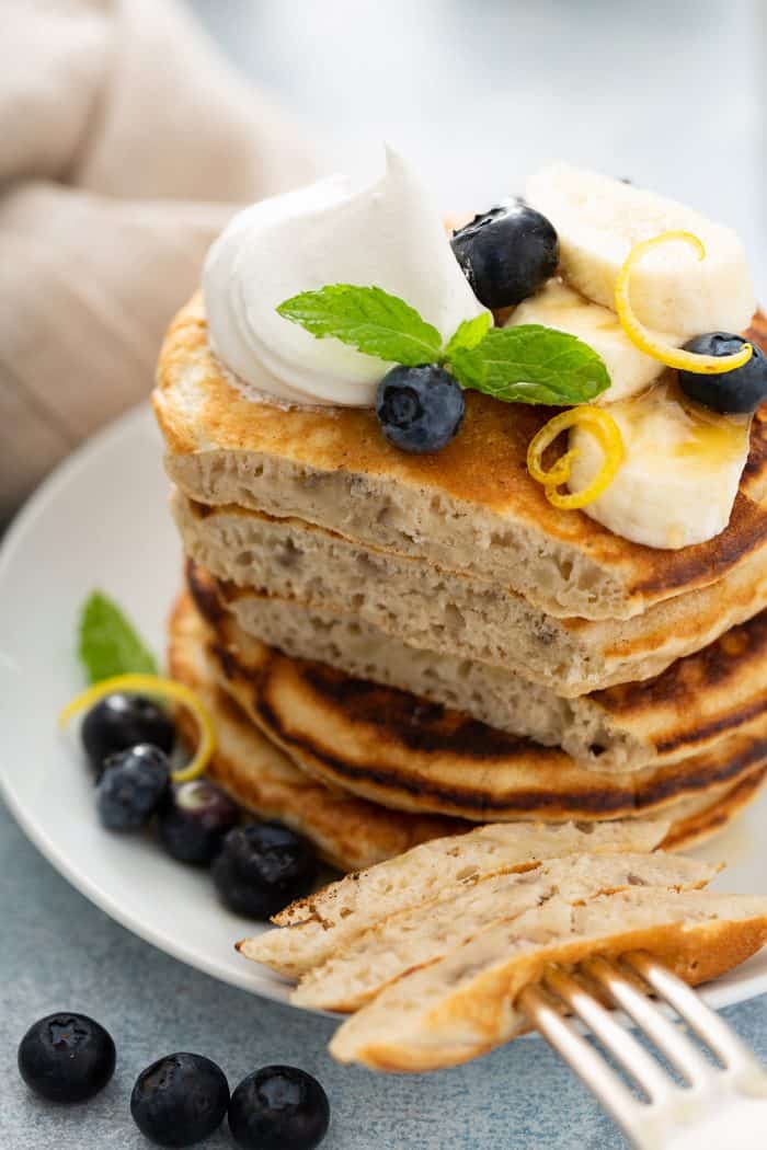 Stack of banana pancakes topped with blueberries, sliced bananas, and whipped cream next to a fork with a bite of pancakes on it