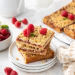 Three raspberry bars stacked on a white plate, with more sliced bars in the background