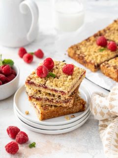 Three raspberry bars stacked on a white plate, with more sliced bars in the background