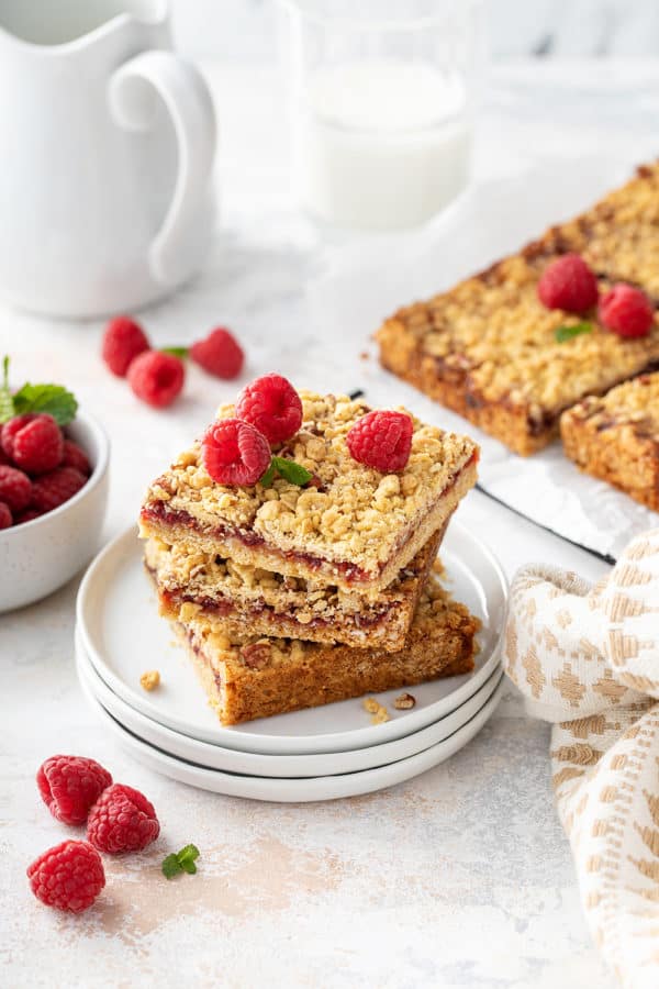 Three raspberry bars stacked on a white plate, with more sliced bars in the background