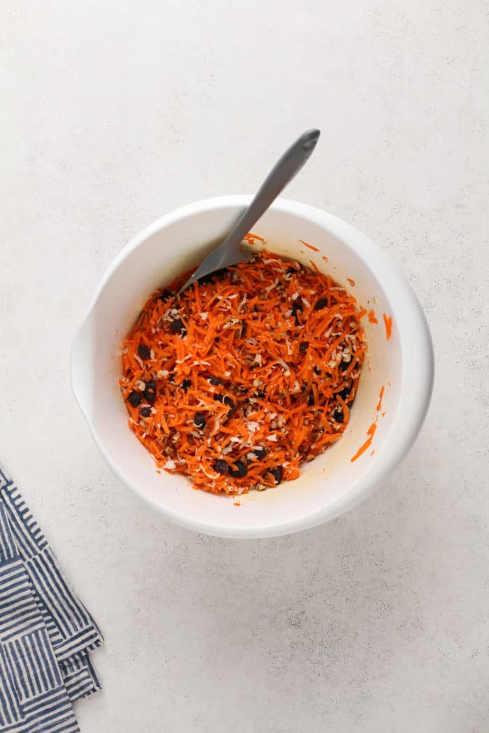 Carrots, nuts, coconut, and raisins mixed in a white mixing bowl.