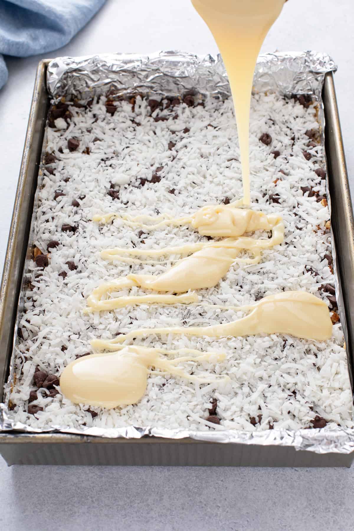 Sweetened condensed milk being poured over coconut layered onto magic bars.
