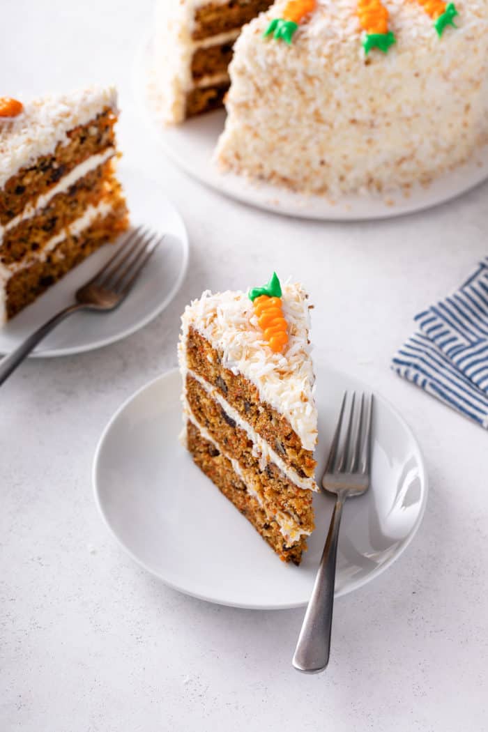 Slice of carrot cake decorated with coconut and an icing carrot next to a fork on a white plate.