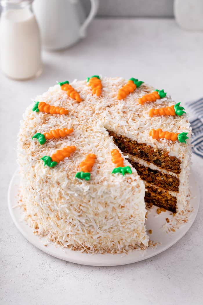Carrot cake covered in coconut and decorated with icing carrots with one slice removed.