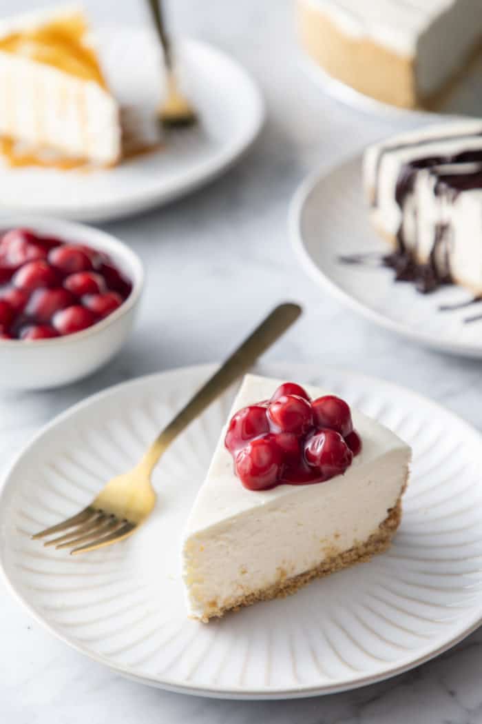No-bake cheesecake topped with cherry pie filling on a white plate, with more slices of cheesecake in the background.