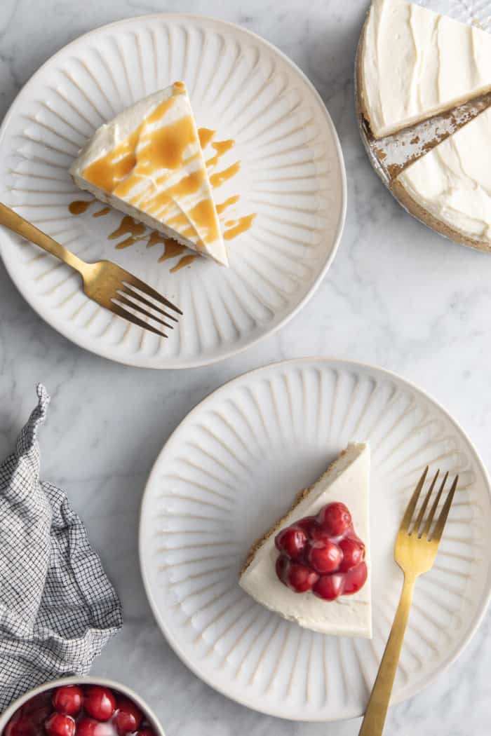 Two white plates with slices of no-bake cheesecake, one topped with caramel sauce and the other topped with cherry pie filling.