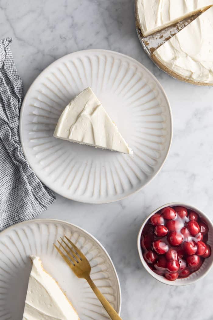 Slices of no-bake cheesecake on white plates next to a bowl of cherry pie filling.