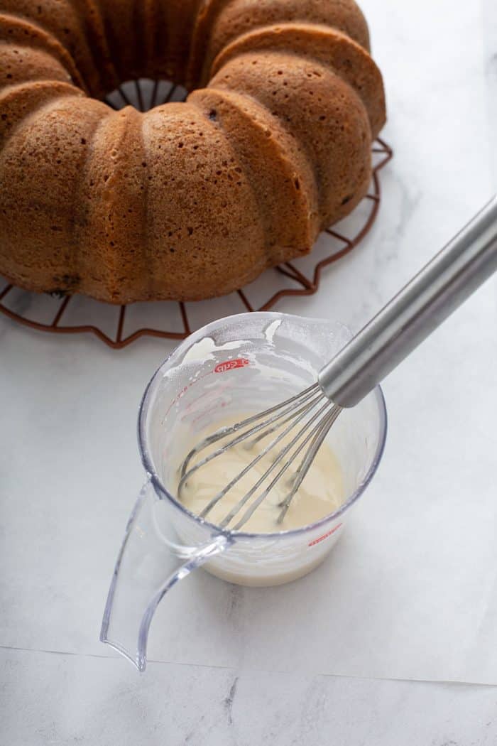 Orange glaze being whisked together in a measuring cup next to a baked bundt cake