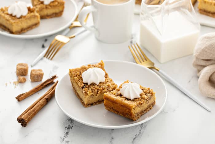 Two pumpkin cheesecake bars on a white plate, with cups of coffee in the background