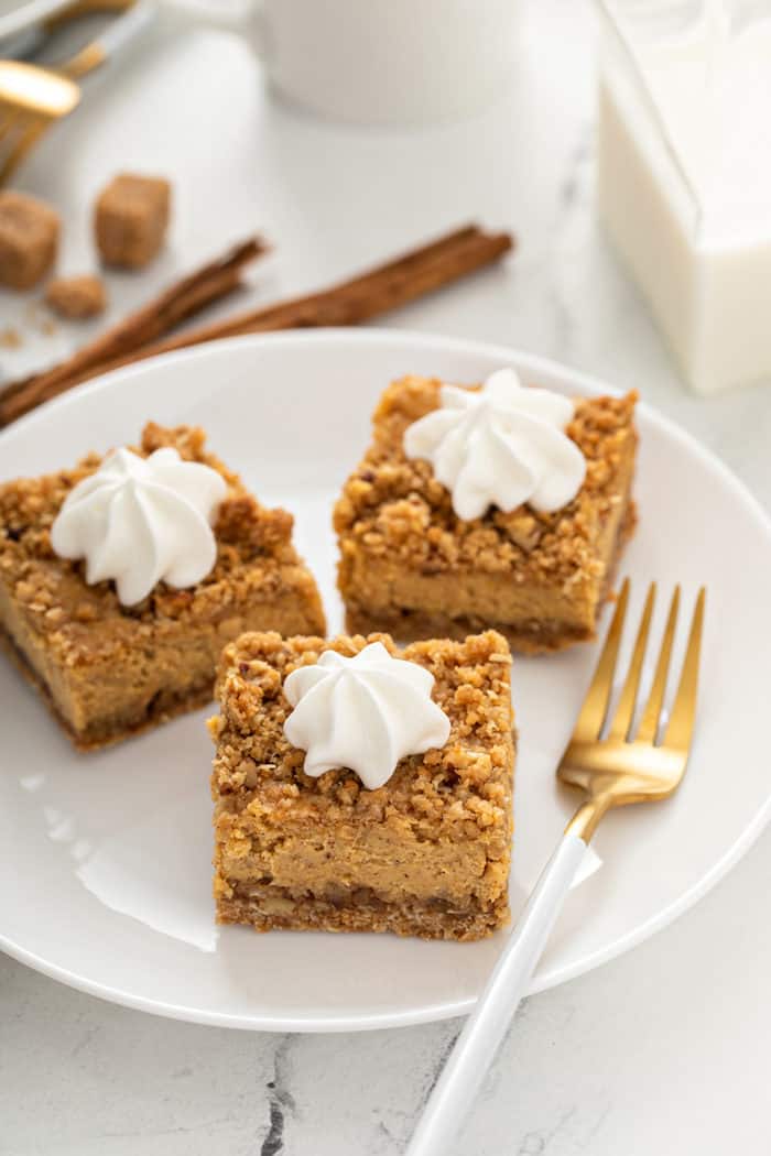 Three pumpkin cheesecake bars, each topped with whipped cream, on a white plate