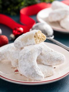 Pile of almond crescent cookies on a plate, with the top cookie broken in half.