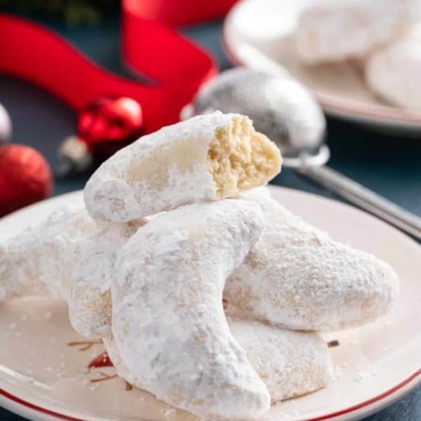 Pile of almond crescent cookies on a plate, with the top cookie broken in half.