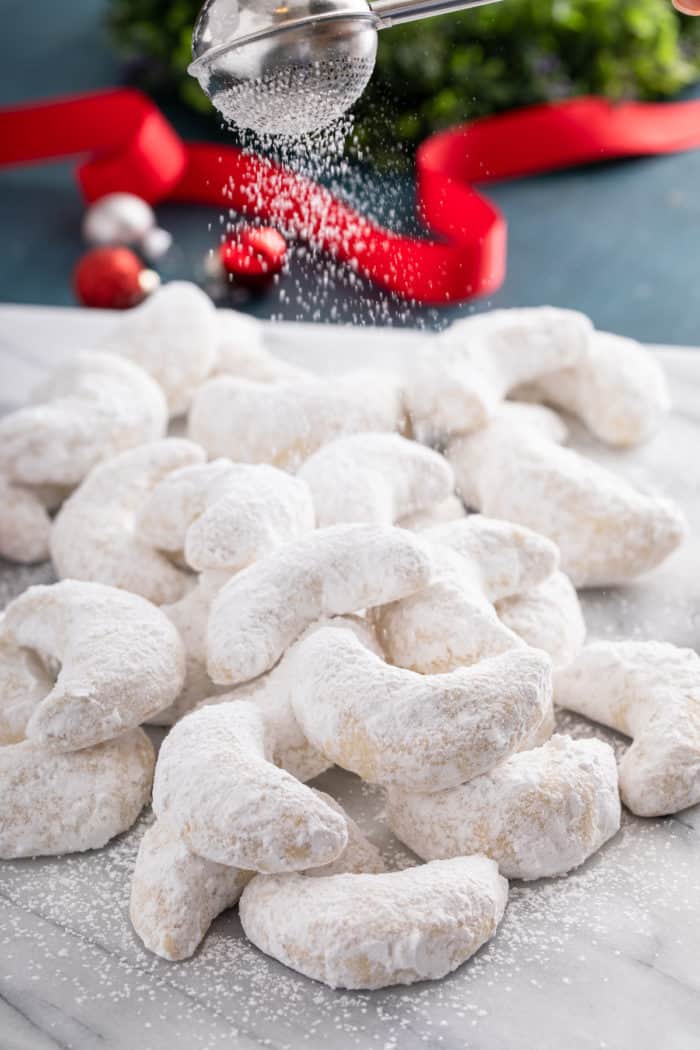 Almond crescent cookies arranged in a pile on a marble board. Powdered sugar is being dusted over the top of the cookies.