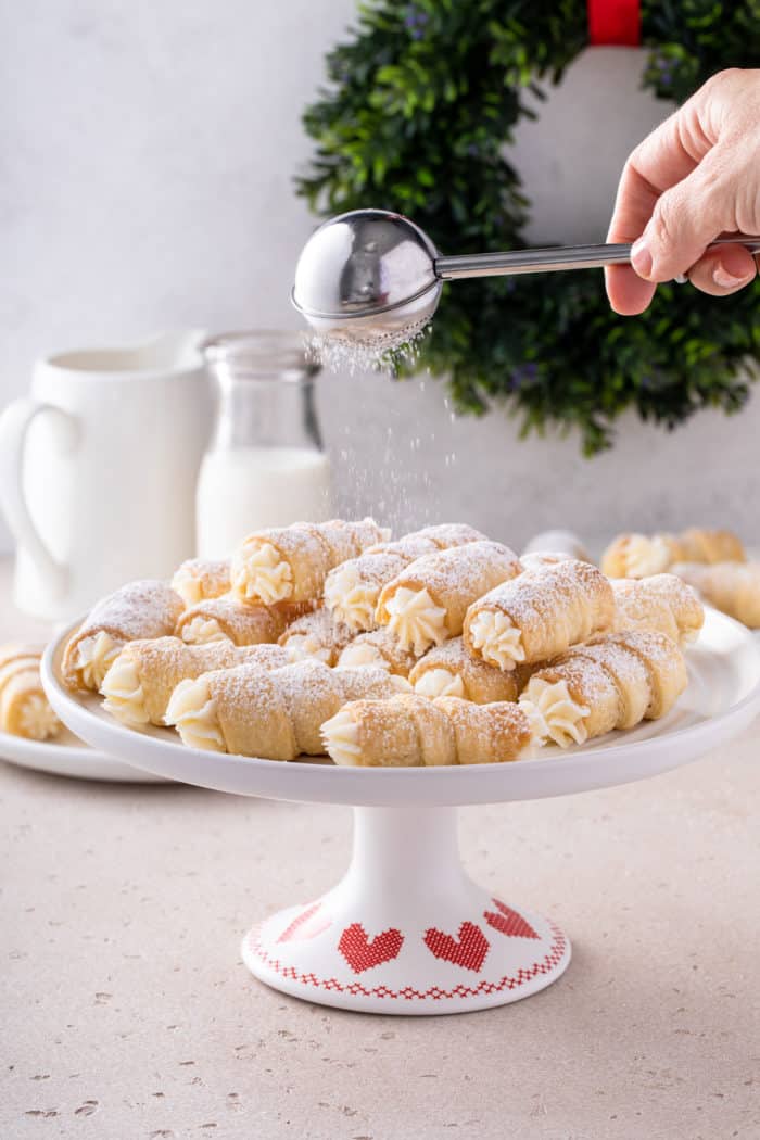 White cake plate piled with clothespin cookies. A hand is dusting powdered sugar over the top of the cookies.